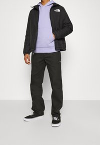 Man in een zwarte North Face-jack, lavendel hoodie, zwarte broek en zwarte sneakers, staand tegen een effen witte achtergrond.