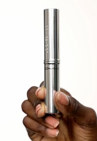Tube de mascara métallique argenté tenu dans une main, présentant une surface lisse, une forme cylindrique et un logo gravé sur le côté.