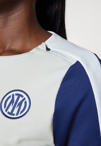 Haut de sport à manches courtes en vert clair et bleu marine, avec un logo brodé, une texture lisse et un détail zippé au col.