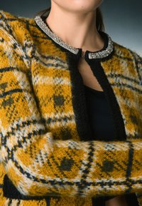 Cárdigan de punto en cuadro amarillo y negro, con un acabado texturizado y un cuello con abalorios. Se abrocha al frente con un pequeño gancho.