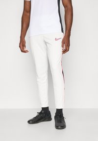 Pantalons de sport blancs avec des rayures roses sur les côtés, associés à un haut blanc et noir à manches courtes, et des chaussures de sport noires.