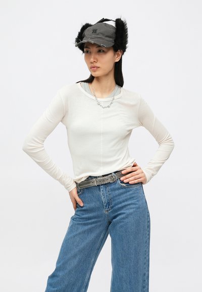 Κρεμ τοπ με μακριά μανίκια, υφασμάτινη υφή· τζιν μπλε ψηλόμεσο· μαύρη ζώνη με ασημένιες τρουκς· γκρι καπέλο με λεπτομέρειες από γούνα.