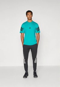 Turkio spalvos trumpomis rankovėmis marškinėliai su juodomis Adidas juostelėmis ant rankovių, derinami su juodomis siaurėjančiomis sportinėmis kelnėmis, kuriose yra baltos Adidas juostelės.