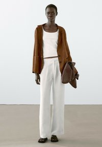 Hnědý sukně blazer, bílý tílko a bílé široké kalhoty s proužky. Drží pletenou hnědou tašku. Celý outfit doplňují černé sandály.