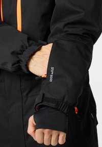 Zwarte jas mouw met elastische manchetten en klittenbandsluiting. Bevat STX™ 8000 merkbranding en een oranje trekband voor aanpassing.