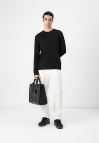 Zwarte lange mouwen shirt, witte broek, zwarte sneakers, houdt een zwarte leren tote bag vast met duidelijke compartimenten en korte handvatten.