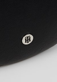 Surface en cuir noir texturé avec un petit emblème métallique argenté rond présentant des lettres entrelacées stylisées.