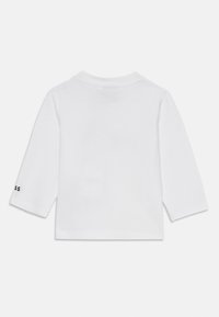 Camisa blanca de manga larga, hecha de algodón, con un cuello redondo y un sutil logo en la manga izquierda en negro. Textura suave, corte casual.