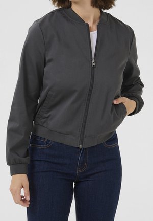 Frau trägt eine dunkelgraue Bomberjacke mit Reißverschluss und gerippten Bündchen sowie blaue Jeans, die Hand in der Jackentasche, vor einem schlichten Hintergrund.