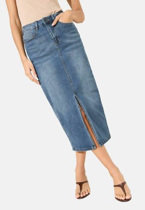 Frau trägt einen blauen Denim-Midirock mit Frontschlitz, helles Oberteil und braune Zehensandalen, steht mit entspannten Händen.