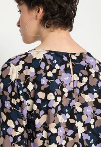 Oberteil mit Blumenmuster in Schwarz, Creme, Lila und Blau. Rückenschluss mit einem kleinen Knopf und einem Schlüsselloch-Detail. Glatte Stofftextur.
