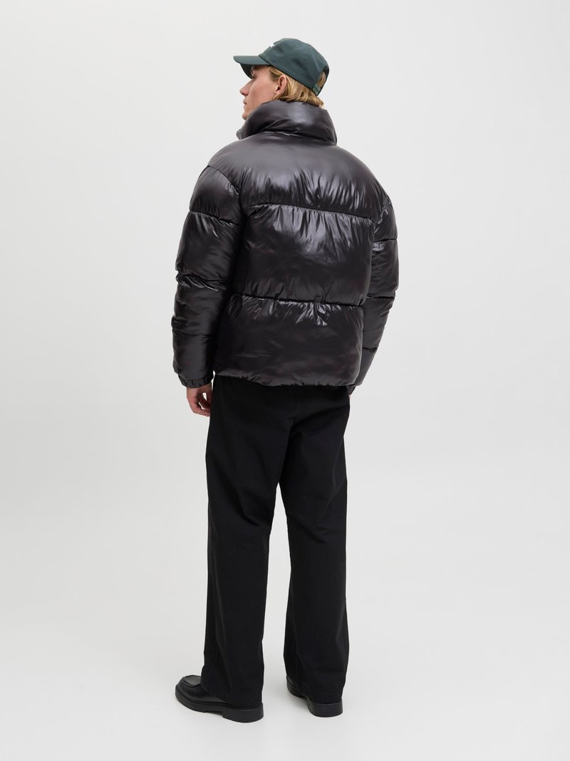 Puffermantel aus glänzendem schwarzem Material, mit hohem Kragen und horizontalem Steppmuster. Kombiniert mit lockeren schwarzen Hosen und schwarzen Schuhen.