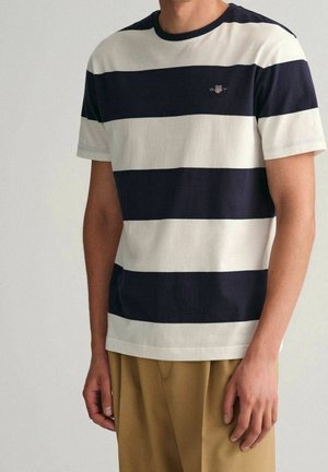 Homme portant un t-shirt à manches courtes avec de larges rayures horizontales bleu marine et blanches et un pantalon beige sur un fond uni.