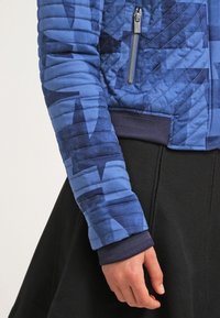 Gepolsterte blaue Jacke mit geometrischem Muster, ausgestattet mit einem silbernen Reißverschluss und gerippten Bündchen. Das Material ist glatt mit unterschiedlicher Textur.