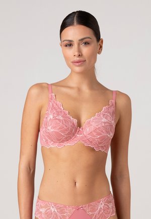 Donna che indossa un reggiseno in pizzo floreale rosa e mutandine a vita alta abbinate con spalline sottili, posando su uno sfondo chiaro uniforme.