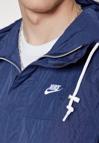 Giacca leggera blu navy con finitura testurizzata, dotata di logo Nike bianco, cappuccio con cordino e zip metallica per la chiusura.