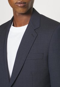 Granatowa marynarka o teksturowanym materiale z klapami w kształcie notcha i pojedynczą kieszenią na piersi, noszona na białej koszulce z okrągłym dekoltem, podkreślająca dopasowany krój kurtki.