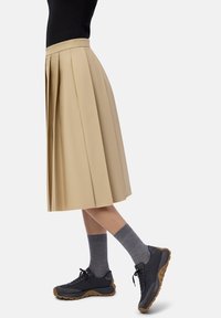 Persona che indossa una gonna midi plissettata beige, calze grigie e sneakers nere con suole beige, in piedi davanti a uno sfondo bianco.