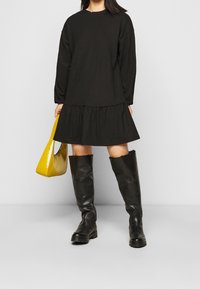 Černé dlouhé šaty s volánkovou sukní, doplněné černými kozačkami po kolena a lesklou žlutou kabelkou. Hladká textura látky.