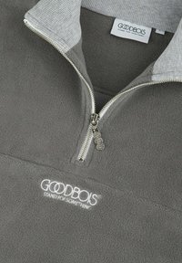 Grauer Fleece-Pullover mit halbem Reißverschluss, geripptem Kragen und gesticktem Logo. Glatte Textur mit dezentem Glanz und silbernen Hardware-Details.