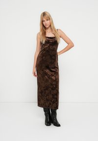 Brązowa aksamitna sukienka maxi z kwiatowym haftem, cienkimi ramiączkami i dopasowaną sylwetką, zestawiona z czarnymi kozakami za kolano.