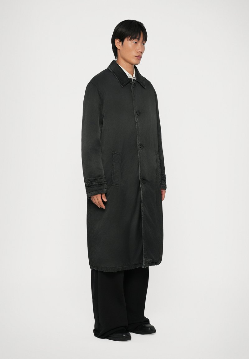 Lungo cappotto nero realizzato in un tessuto liscio, con chiusura a bottoni, due tasche laterali e un colletto strutturato. Vestibilità leggermente oversize.