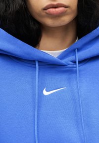 Κοντινό πλάνο ενός ατόμου που φοράει μπλε φούτερ με κουκούλα Nike, με ορατό λευκό λογότυπο και κορδόνια κουκούλας, εμφανίζοντας τα χείλη και τον λαιμό.