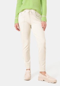 Cream-colored trousers with a slim fit, featuring an elastic waistband, stitch detailing, and side pockets, paired with light beige shoes.