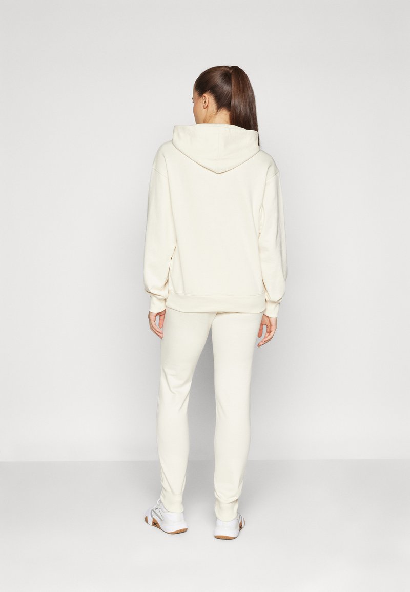Sudadera crema con capucha y mangas largas, combinada con pantalones ajustados a juego. Tejido suave y corte relajado; exhibida por la parte de atrás.