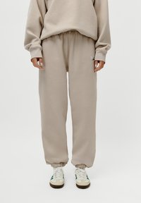 Beige hettegenser og joggebukse sett i myk stoff, med elastiske mansjetter og midjebånd. Sko med grønne og blå striper fullfører antrekket.