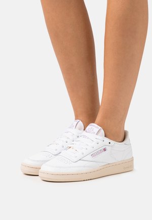 Zapatillas de cuero blanco con un diseño de corte bajo, que presentan una suela de goma beige claro, acentos del logo en púrpura y detalles perforados en la puntera.