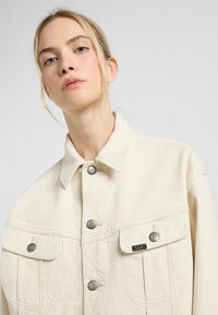Beige farkkutakki, jossa on kauluspanta, hopeiset nepparit ja kaksi etutaskua. Takissa on hienovarainen logopatch rintataskun lähellä.