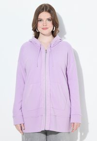 Veste à zip lavande avec de grandes poches avant, capuche avec cordon de serrage et coupe relaxée. Le tissu est lisse avec une légère texture.