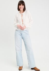 Blouse blanche en broderie anglaise avec des manches bouffantes et des liens sur le devant, associée à un jean large bleu clair et des bottines brunes.