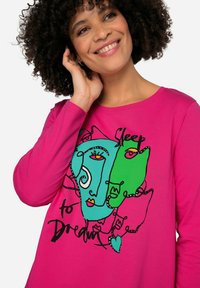 Camisa de manga larga fucsia con diseño de cara abstracta en verde y azul, con contornos negros y el texto "Dormir para soñar."