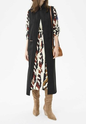 Kobieta ubrana w sukienkę w wzory z guzikami, długą czarną kamizelkę bez rękawów, beżowe zamszowe kozaki za kolano oraz niosąca brązową torebkę na ramię.