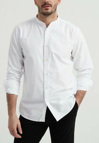Chemise blanche à manches longues avec un col montant, une fermeture à boutons, et des poignets retroussés, associée à un pantalon noir.
