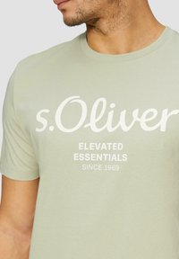 Hele roheline puuvillane t-särk, millel on ümar krae, lühikesed varrukad ja valge tekst: "s.Oliver", "ELEVATED ESSENTIALS", "SINCE 1969".