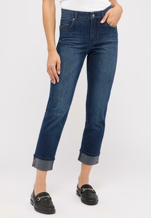 Jean en denim bleu foncé, taille haute, coupe droite, avec des poignets roulés mettant en valeur une doublure bleu clair contrastante. Dispose de poches avant.