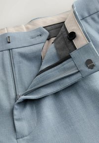 Gros plan sur un pantalon bleu clair sur mesure avec fermeture éclair, crochet et agrafe, ainsi qu'un bouton noir, montrant la doublure intérieure et les détails de la ceinture.