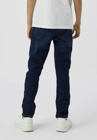 Jeans de mezclilla de corte slim en azul oscuro, con bolsillos traseros y costuras en contraste, combinados con zapatillas blancas, vistos desde la parte trasera.