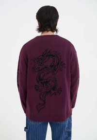 Burgundy pletený svetr s černým designem draka na zadní straně, s žebrovanými rukávy a lemem, doplněný modrými pruhovanými džíny.