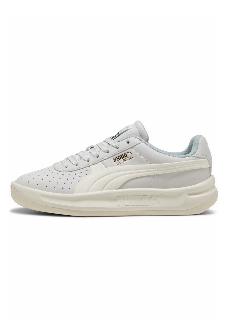 Zapatilla blanca con diseño perforado, acentos en azul claro y suela texturizada. Presenta un logo dorado en el costado y cordones planos.
