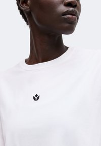 Gros plan d'une personne portant un t-shirt blanc uni avec un petit logo fleur abstrait noir près de l'encolure.