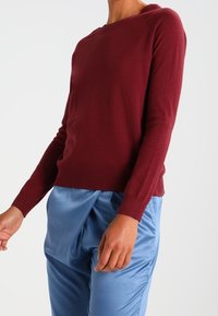 Pull en maille ajusté couleur bordeaux avec un large col rond et des manches longues, associé à un pantalon bleu clair drapé. Texture lisse.
