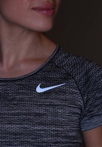 Camisa deportiva de patrón negro y gris con cuello redondo y mangas cortas, con un logo blanco de Nike en el pecho.