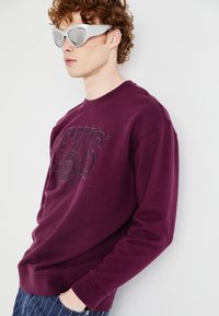 Felpa bordeaux con logo in rilievo sul davanti, collo rotondo e polsini a costine. Indossata con occhiali da sole grigi futuristici e pantaloni di denim.