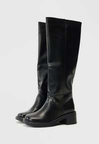 Bottes en cuir noir hautes avec un embout arrondi, un talon épais et une texture lisse. Le design présente une silhouette propre et minimaliste.