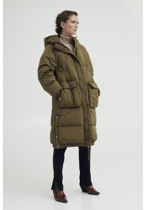 Olijfgroene pufferjas met een capuchon, met grote voorzakken, een getailleerde taille en drukknoopsluitingen. Gekozen met zwarte flare jeans en bruine hakken.