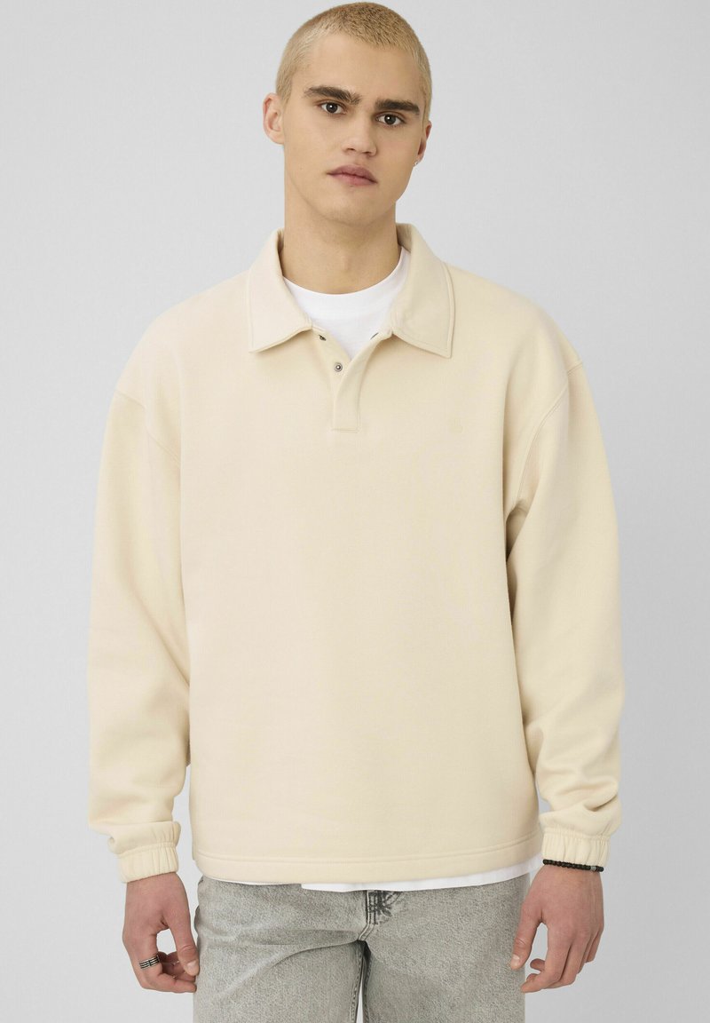 Jeune homme aux cheveux blonds courts portant un pull beige à col, une chemise intérieure blanche, un jean clair, debout devant un fond uni.